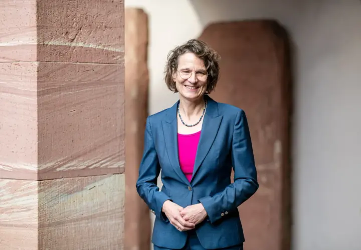 Christiane Tietz, eine Frau mit Brille und lockigem Haar, lächelt in einem blauen Hosenanzug vor einer Sandsteinwand.
