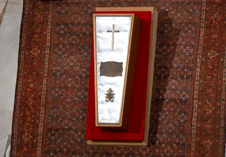 Der gläserne Sarg eines Papstes, mit Kreuz und Wappen, liegt auf einem roten Samtkissen vor einem reich gemusterten Teppich im Petersdom.