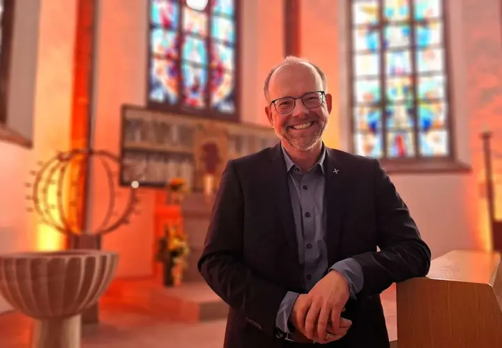 Lächelnder Mann in Anzug lehnt an einer Kirchenbank. Buntglasfenster und Taufbecken im Hintergrund. Hier kann man wieder in die Kirche eintreten.