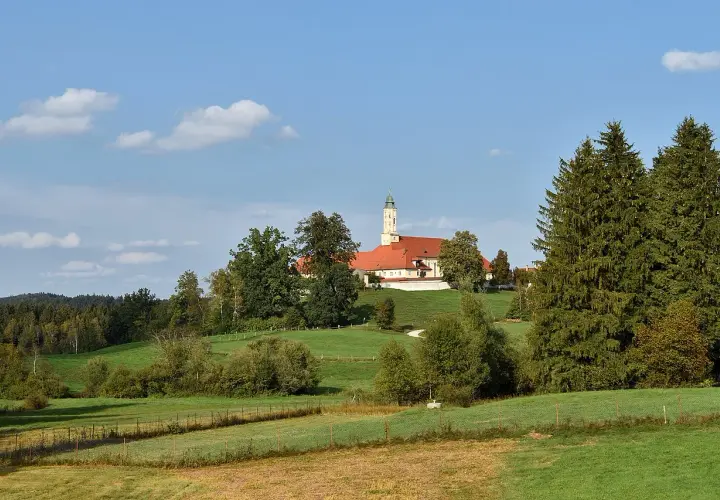 Kloster Reutberg thront auf einem Hügel, umgeben von grünen Wiesen und Bäumen unter blauem Himmel mit Wolken.