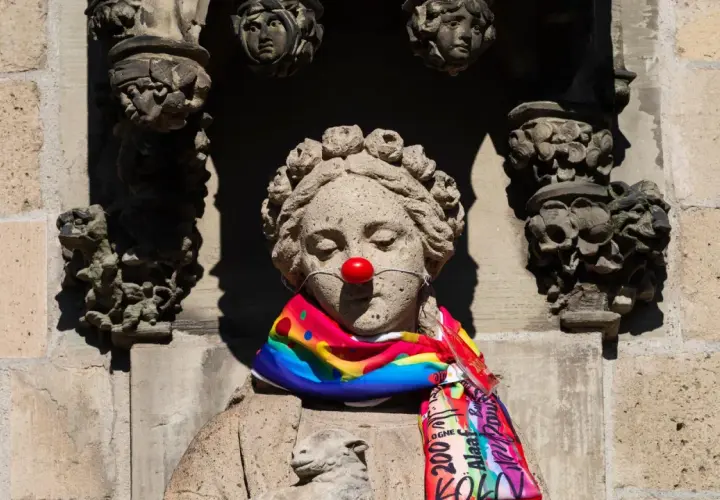 Steinskulptur mit roter Clownsnase und buntem Schal. Die Frage "was hat Karneval mit Religion zu tun?" wird hier humorvoll aufgeworfen.