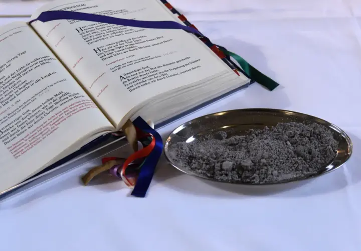Ein aufgeschlagenes Buch mit Gebeten und eine Schale mit Asche. "Aus Staub bist du zu Staub wirst du" – ein Symbol für Vergänglichkeit.