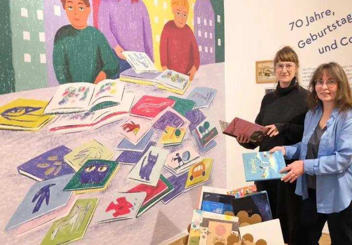 Zwei Frauen stehen vor einer bunten Wandmalerei mit Büchern und halten Bücher in den Händen.