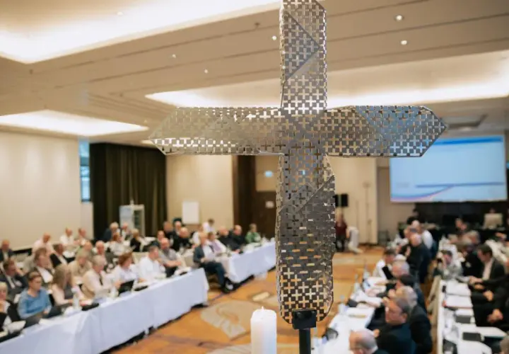 Ein moderner Kreuz-Schriftzug dominiert die Szene, während im Hintergrund ein synodaler Ausschuss tagt.