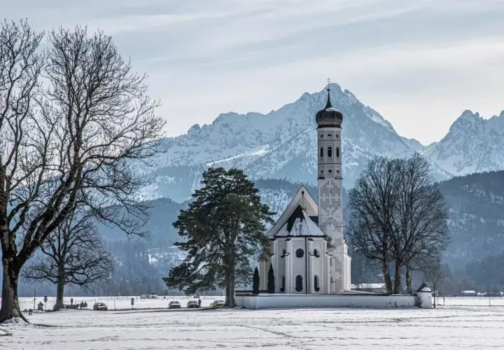 Winterliche Szene mit der Kirche St. Coloman im bayerischen Voralpenland, schneebedeckte Berge im Hintergrund.