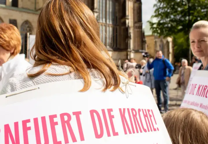 Frauen mit Schildern fordern "Erneuert die Kirche!". Eine Frau mit roten Haaren und eine mit blonden Haaren sind zu sehen.