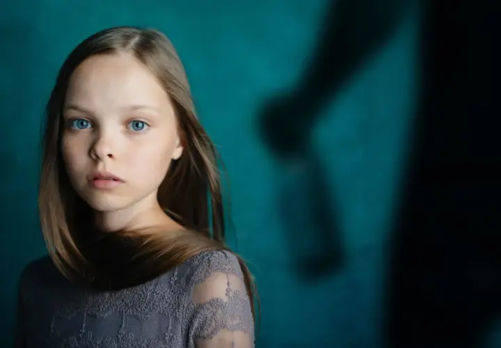 Ein junges Mädchen mit blauen Augen blickt nachdenklich, während im Hintergrund eine schattenhafte Figur erscheint, die an einen beschützenden Gott als Vater erinnert.
