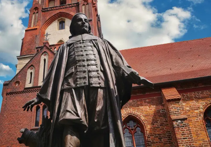 Bronzestatue von Paul Gerhardt vor einer roten Backsteinkirche mit blauen Himmel und Wolken.