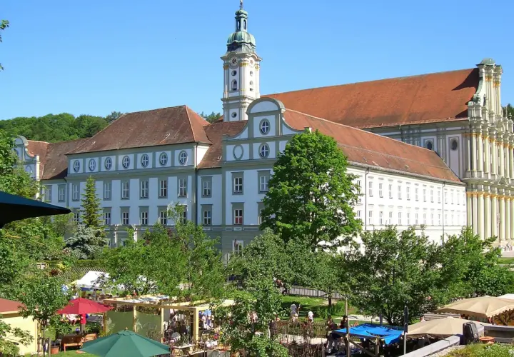 Prächtiges Kloster FFB mit barockem Turm, umgeben von grünen Bäumen und einem lebhaften Markt im Vordergrund.