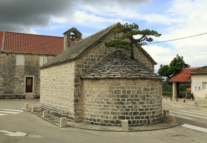 Kleine Steinkirche mit halbrunder Altarnische, bedeckt mit Schindeln. Ein Baum wächst aus dem Dach.