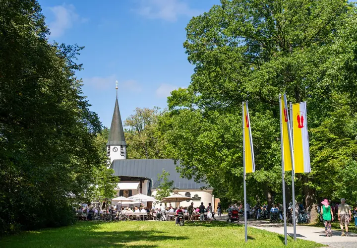 Ein sonniger Tag bei Maria Eich Planegg. Menschen sitzen unter Sonnenschirmen vor der Kirche, während Fahnen im Wind wehen.