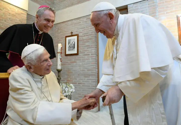Papst Franziskus und ein Bischof lächeln, während sie Benedikt XVI. die Hand schütteln.
