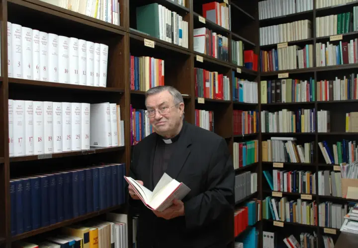 Kardinal Lehmann steht in einer Bibliothek und hält ein Buch in den Händen. Um ihn herum sind Regale voller Bücher.