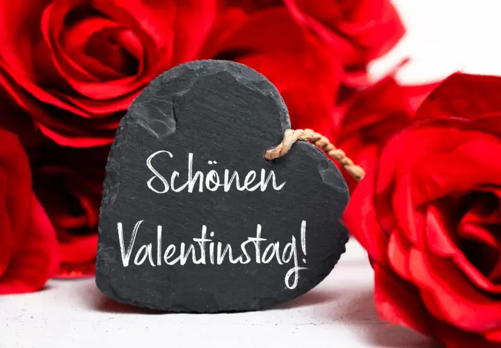 Herzförmiges Schild mit der Aufschrift "Schönen Valentinstag!" vor roten Rosen. Ein liebevoller Gruß zum heiligen Valentin.