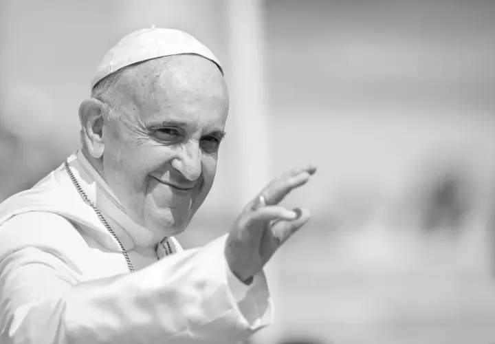 Papst Franziskus winkt lächelnd in die Menge, gekleidet in seine Soutane und mit einer Kette.