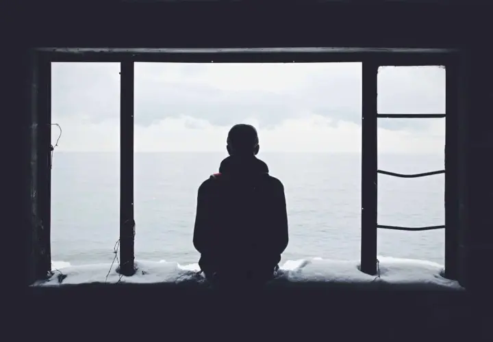 Eine Person blickt aus einem Fenster auf das Meer. Schnee liegt auf dem Sims. Das Bild vermittelt ein Gefühl von Einsamkeit.