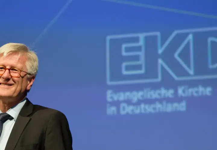 Lächeln des EKD Vorsitzenden vor dem Logo der Evangelischen Kirche in Deutschland.