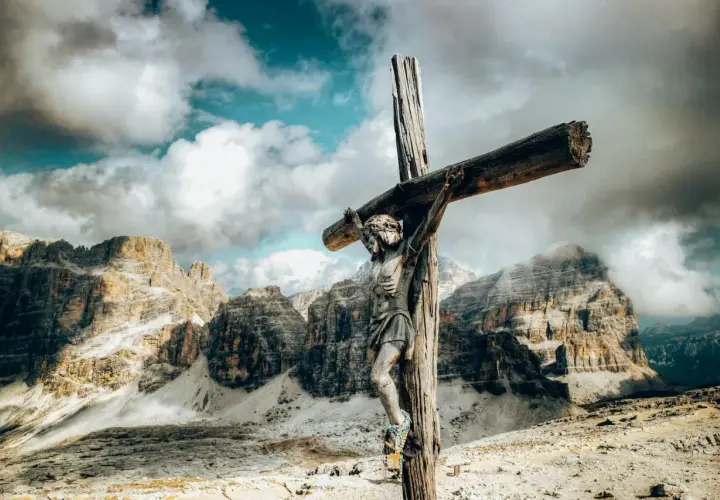 Holzfigur der Jesus-Kreuzigung vor dramatischer Bergkulisse und Wolkenhimmel.