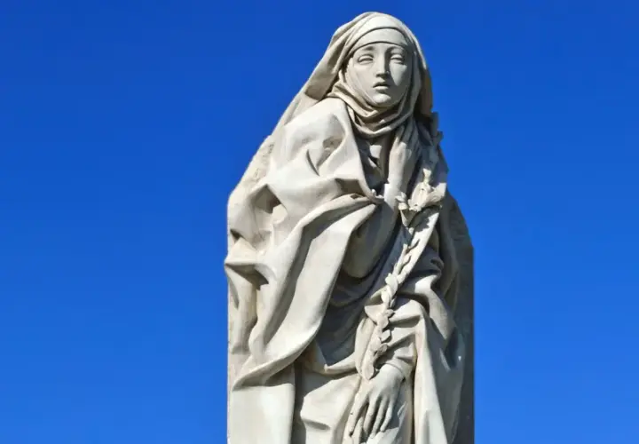 Steinskulptur der Heiligen Katharina von Siena mit Schleier und einem Lorbeerkranz vor blauem Himmel.