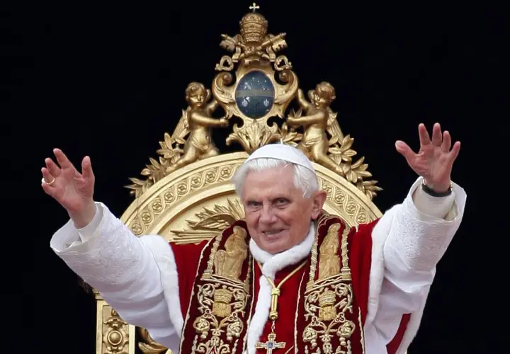 Papst Benedikt XVI. winkt in prächtiger Papst-Kleidung, mit weißer Soutane und rotem Umhang, verziert mit goldenen Stickereien.