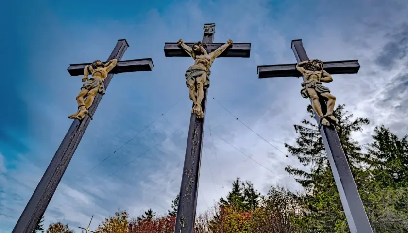 Drei Kreuze mit Jesusfiguren, die die Passion Christi darstellen, vor einem dramatischen Himmel und herbstlichem Laub.