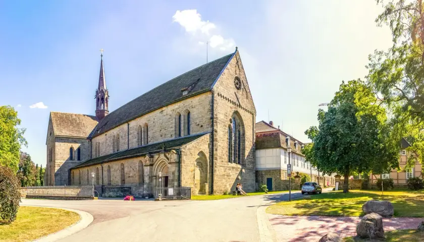 Klosterkirche mit Turm, Teil des Jakobsweg Hessen. Ein Paar sitzt auf einer Bank im Schatten eines Baumes.