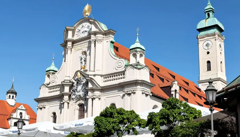 Prachtvolle Kirche am Viktualienmarkt mit goldenem Stern und Glockenturm. Sonnige Szene mit Cafés und Grünflächen.