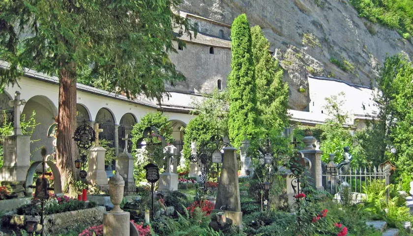 Gräberfeld mit Kreuzen und Urnen vor Arkaden der Erzabtei Salzburg. Üppige Vegetation und Felswand im Hintergrund.