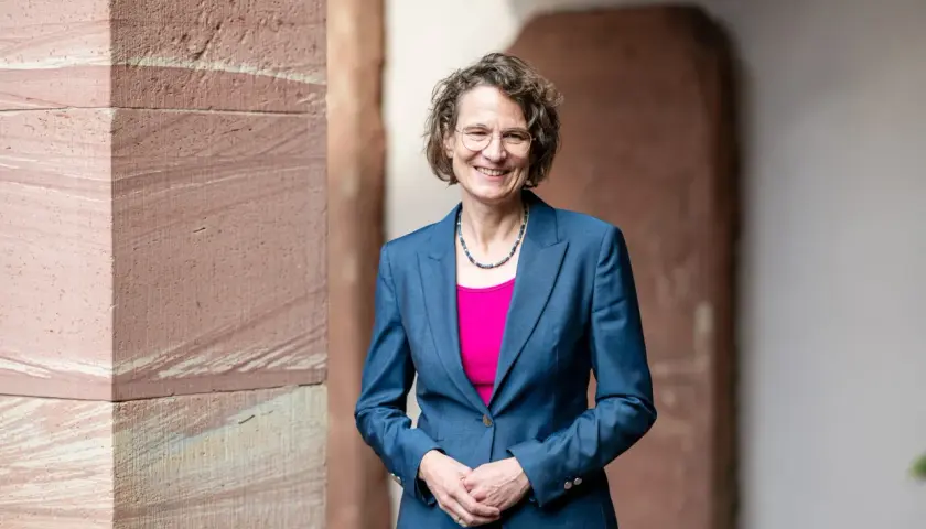 Christiane Tietz, eine Frau mit Brille und lockigem Haar, lächelt in einem blauen Hosenanzug vor einer Sandsteinwand.
