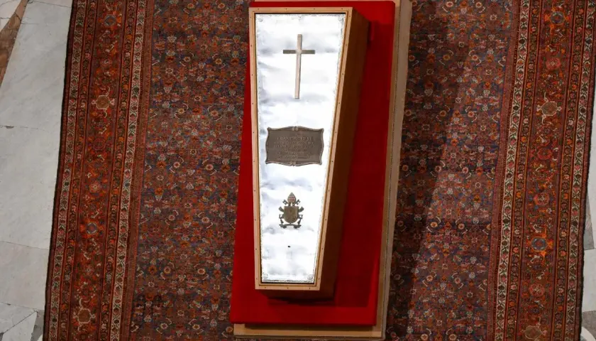 Der gläserne Sarg eines Papstes, mit Kreuz und Wappen, liegt auf einem roten Samtkissen vor einem reich gemusterten Teppich im Petersdom.