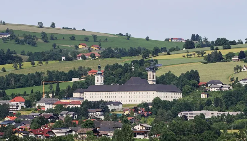 Das Stift Schlierbach, ein imposantes Barockgebäude, thront über einer malerischen Landschaft mit grünen Hügeln und traditionellen Häusern.