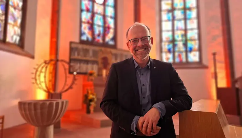 Lächelnder Mann in Anzug lehnt an einer Kirchenbank. Buntglasfenster und Taufbecken im Hintergrund. Hier kann man wieder in die Kirche eintreten.