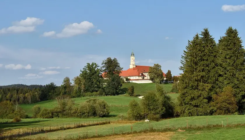 Kloster Reutberg thront auf einem Hügel, umgeben von grünen Wiesen und Bäumen unter blauem Himmel mit Wolken.