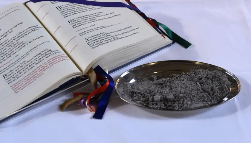 Ein aufgeschlagenes Buch mit Gebeten und eine Schale mit Asche. "Aus Staub bist du zu Staub wirst du" – ein Symbol für Vergänglichkeit.