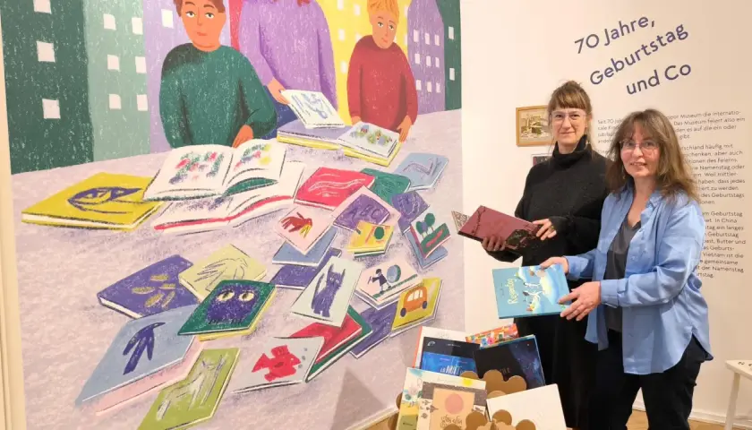 Zwei Frauen stehen vor einer bunten Wandmalerei mit Büchern und halten Bücher in den Händen.