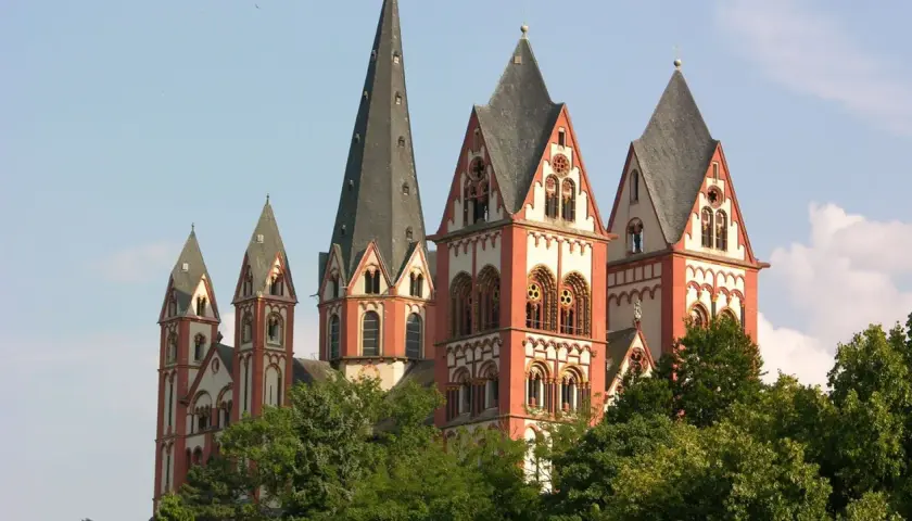 Der Limburger Dom mit seinen roten Türmen ragt über grünen Bäumen auf. Man munkelt, der Bischof von Limburg hätte dort eine goldene Badewanne versteckt.