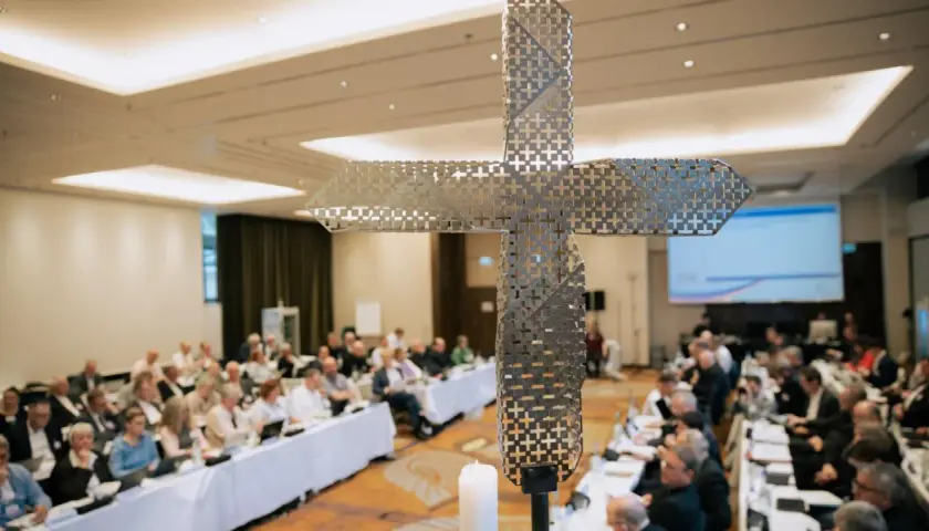 Ein moderner Kreuz-Schriftzug dominiert die Szene, während im Hintergrund ein synodaler Ausschuss tagt.