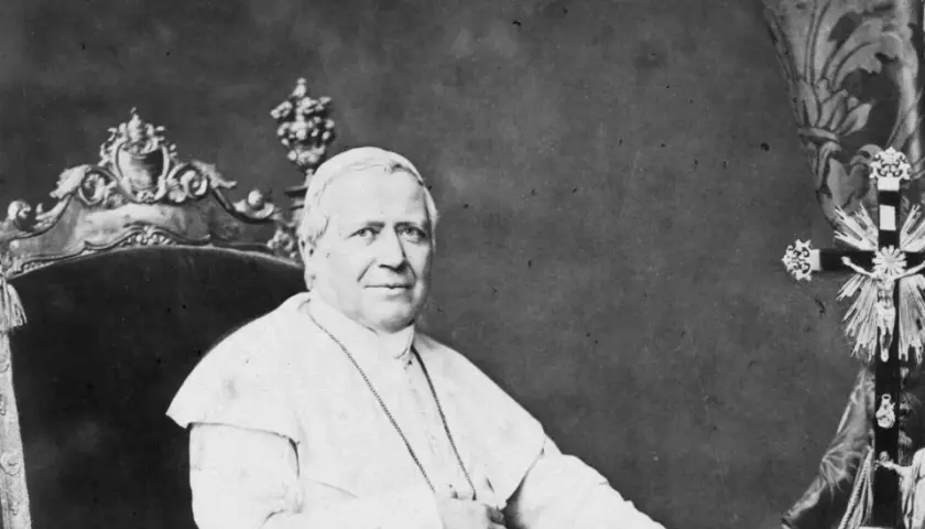 Papst Pius sitzt auf einem prunkvollen Thron, gekleidet in seine Soutane. Neben ihm steht ein aufwendig verziertes Kreuz.