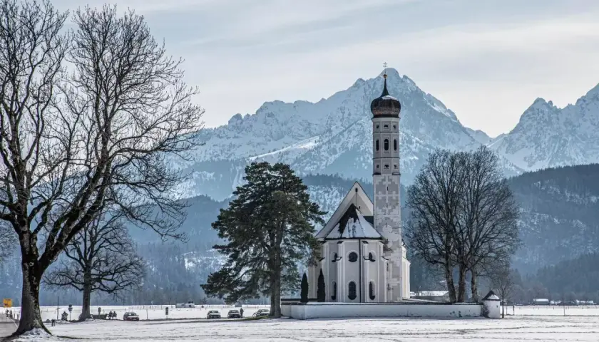 Winterliche Szene mit der Kirche St. Coloman im bayerischen Voralpenland, schneebedeckte Berge im Hintergrund.