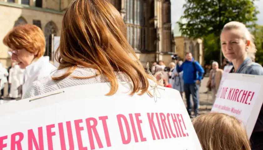 Frauen mit Schildern fordern "Erneuert die Kirche!". Eine Frau mit roten Haaren und eine mit blonden Haaren sind zu sehen.