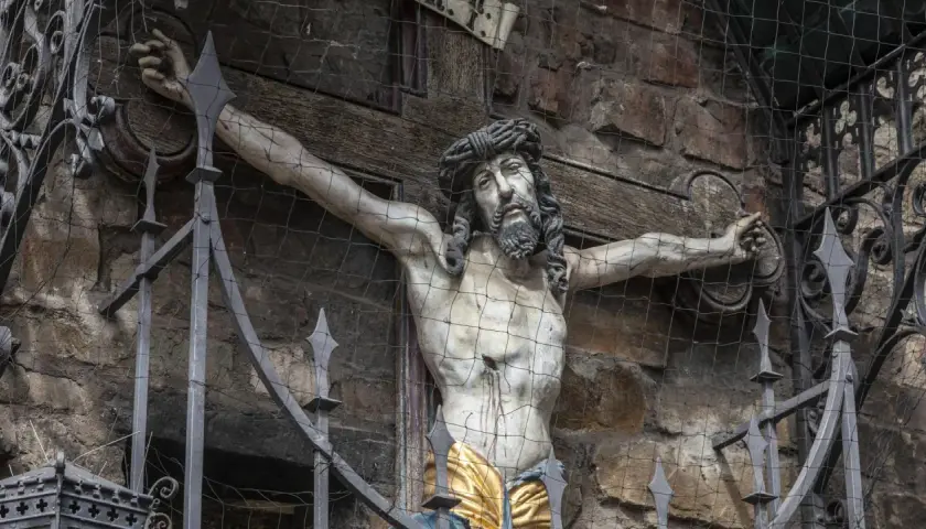 Ein detailreiches, altes jesus kreuz original, hinter einem Metallgitter und Netz, zeigt die leidende Figur Christi vor einer Steinwand.