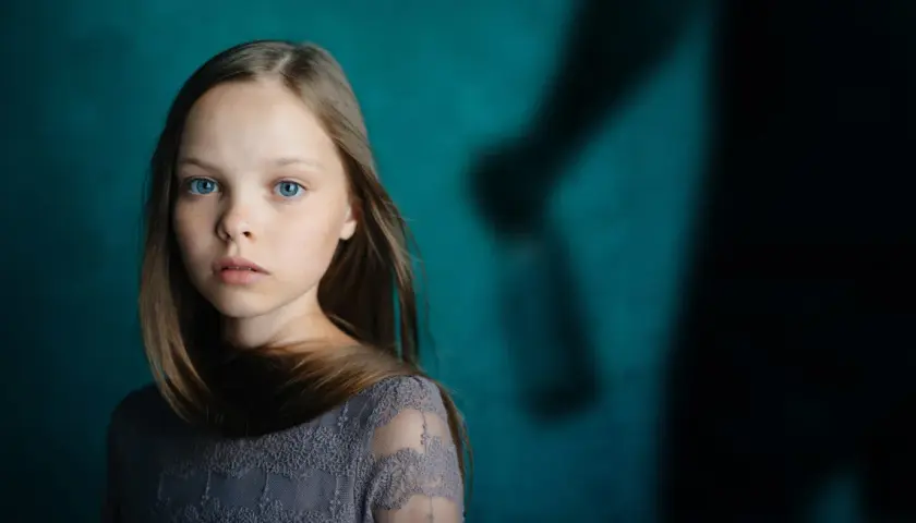 Ein junges Mädchen mit blauen Augen blickt nachdenklich, während im Hintergrund eine schattenhafte Figur erscheint, die an einen beschützenden Gott als Vater erinnert.