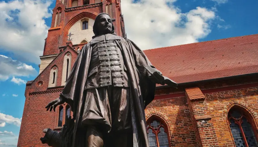 Bronzestatue von Paul Gerhardt vor einer roten Backsteinkirche mit blauen Himmel und Wolken.