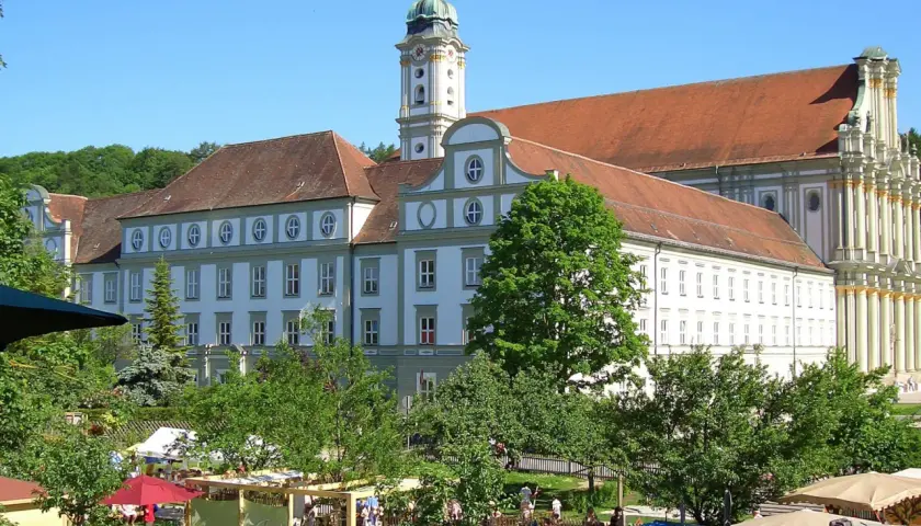 Prächtiges Kloster FFB mit barockem Turm, umgeben von grünen Bäumen und einem lebhaften Markt im Vordergrund.