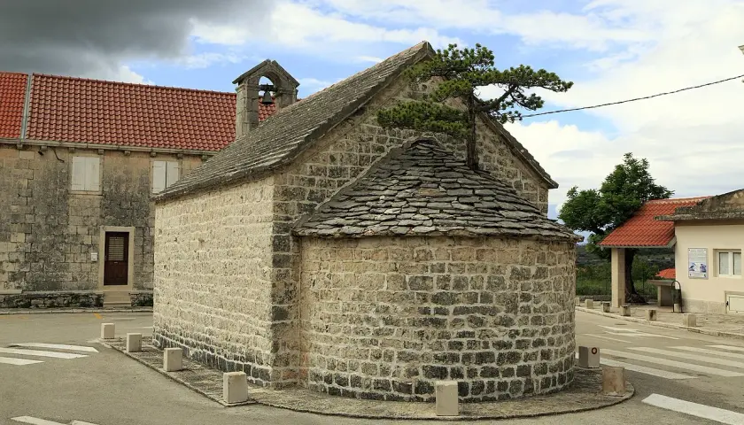 Kleine Steinkirche mit halbrunder Altarnische, bedeckt mit Schindeln. Ein Baum wächst aus dem Dach.