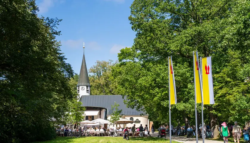 Ein sonniger Tag bei Maria Eich Planegg. Menschen sitzen unter Sonnenschirmen vor der Kirche, während Fahnen im Wind wehen.