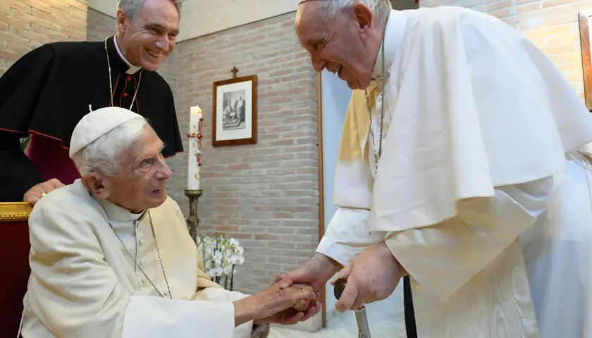 Papst Franziskus und ein Bischof lächeln, während sie Benedikt XVI. die Hand schütteln.