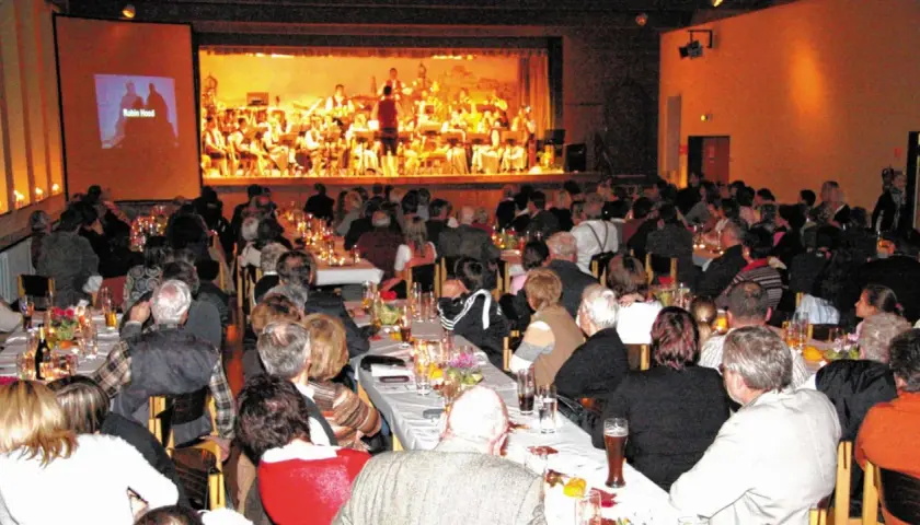 Festliches Bankett im bischof bernhard haus kraiburg. Gäste lauschen einem Orchester, während auf der Leinwand "Robin Hood" läuft.