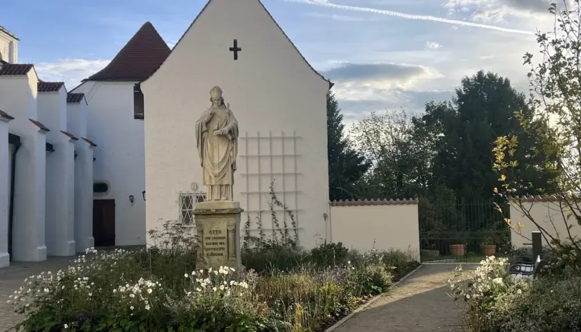 Statue vor dem Domberg Freising. Ein Weg führt durch einen blühenden Garten zu einem Tor.