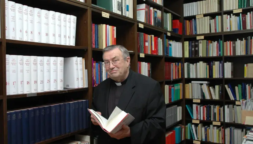 Kardinal Lehmann steht in einer Bibliothek und hält ein Buch in den Händen. Um ihn herum sind Regale voller Bücher.